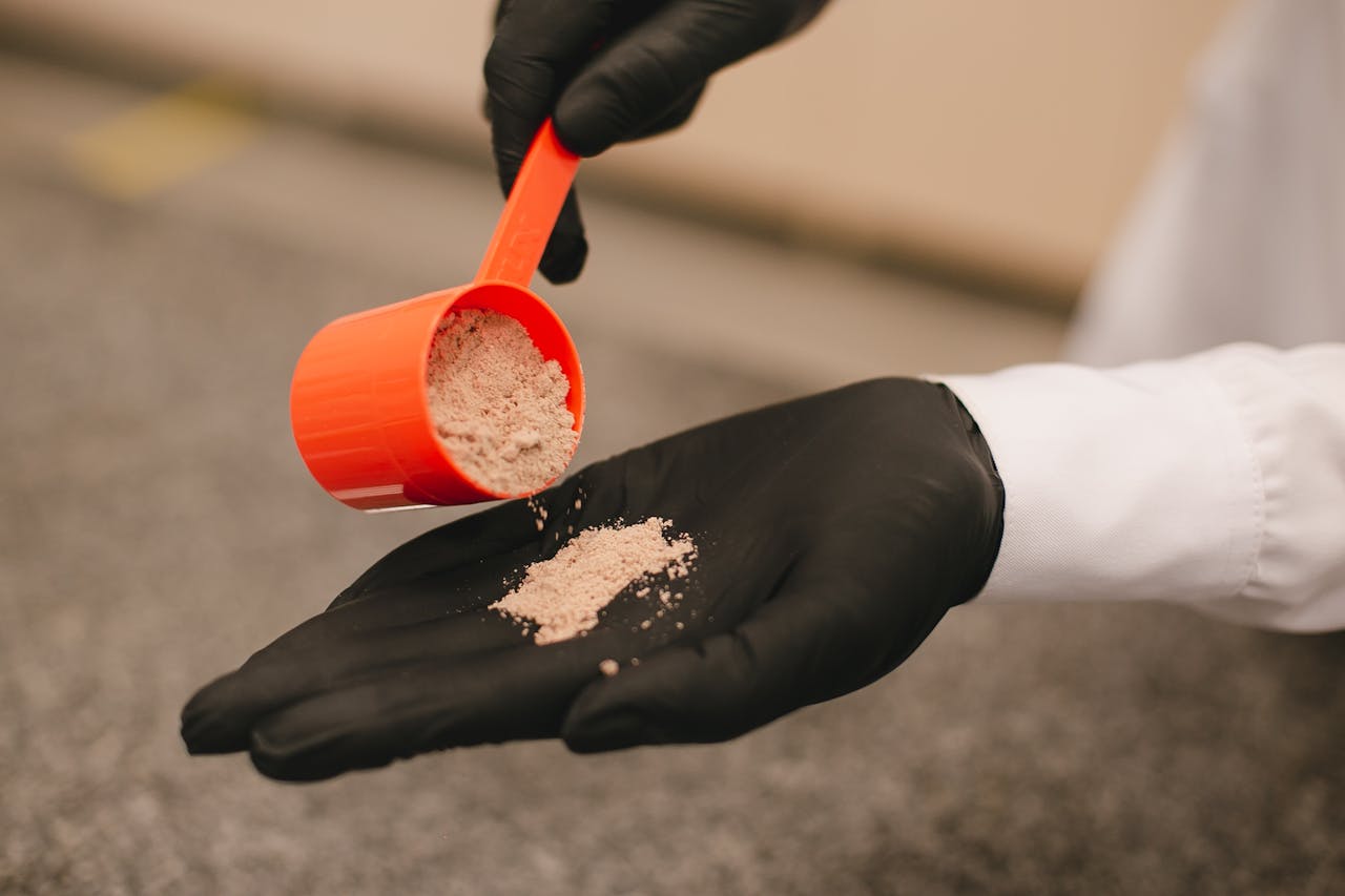 A close-up shot of a black-gloved hand pouring powder from a red scoop, over a textured surface.